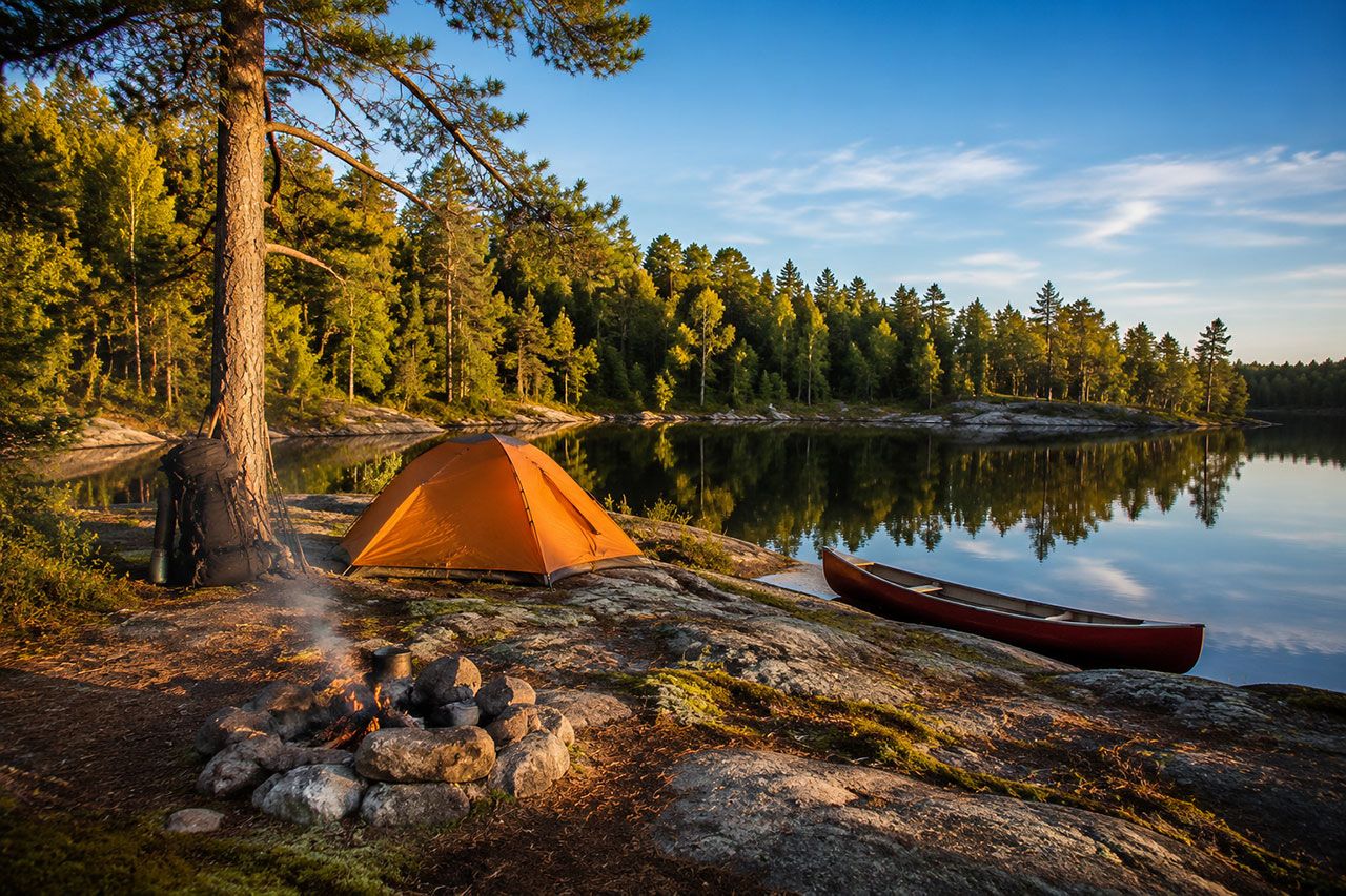 backcountry campsite setup on rocky Canadian Shield terrain with orange tent pitched near pristine lake shoreline. Backpack leaning against white pine tree, campfire ring with stones in foreground, canoe pulled onto shore. Late afternoon golden hour lighting, clear blue sky reflected in calm water. Dense boreal forest backdrop with mix of coniferous and deciduous trees. Authentic wilderness camping scene showing proper Leave No Trace setup, professional outdoor magazine quality.